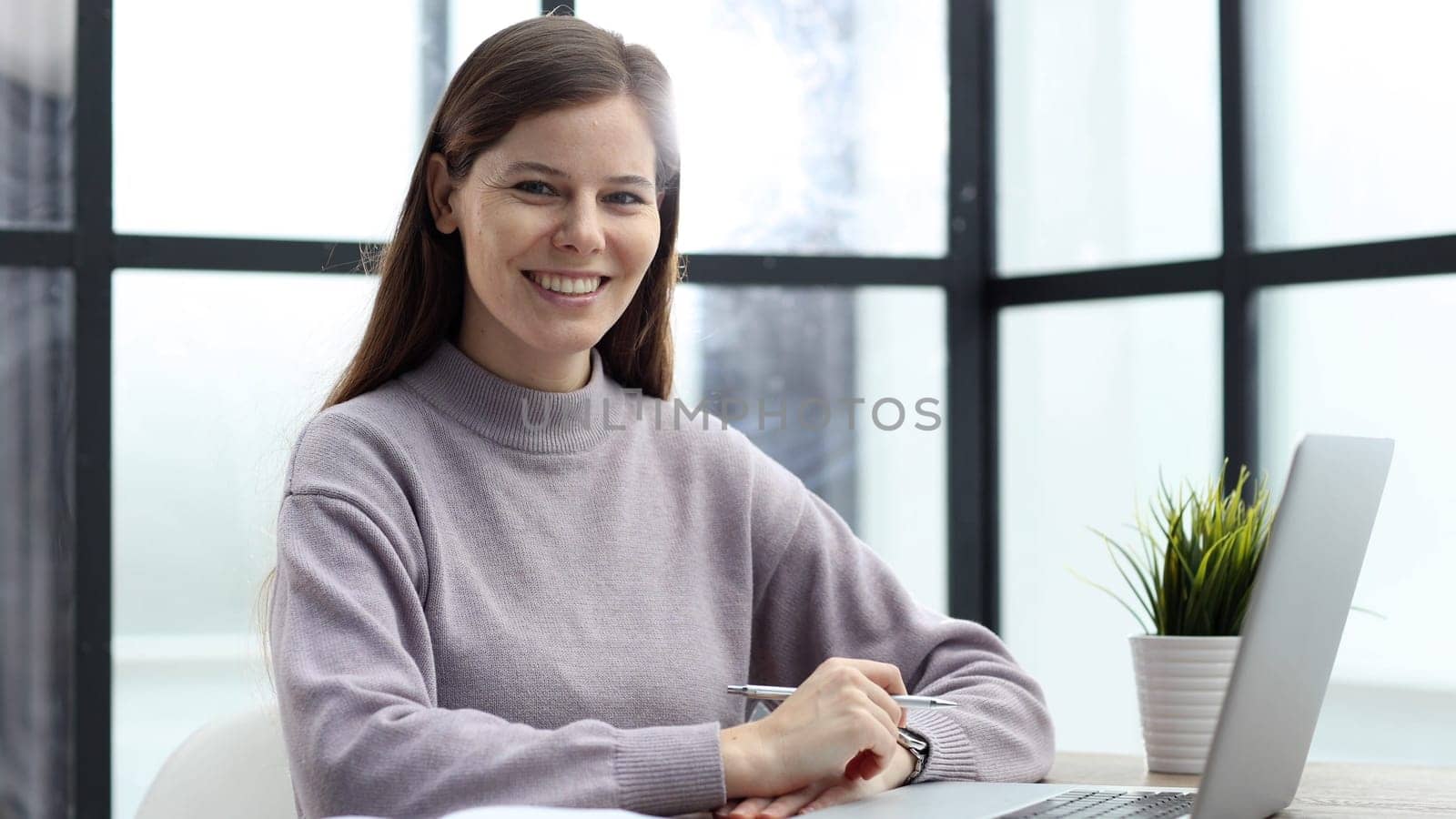 A woman in close-up at a desk in the office working and looking at the camera with a smile.
