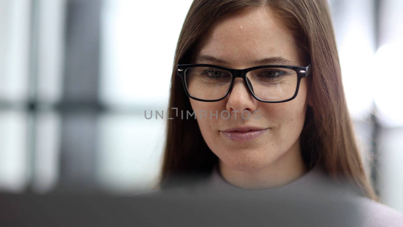A woman with glasses in close-up working at a desk in the office on a laptop.