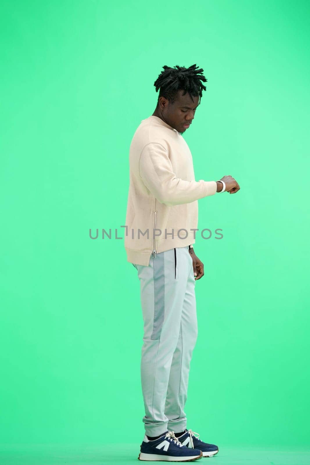 A man, full-length, on a green background, looks at his watch.