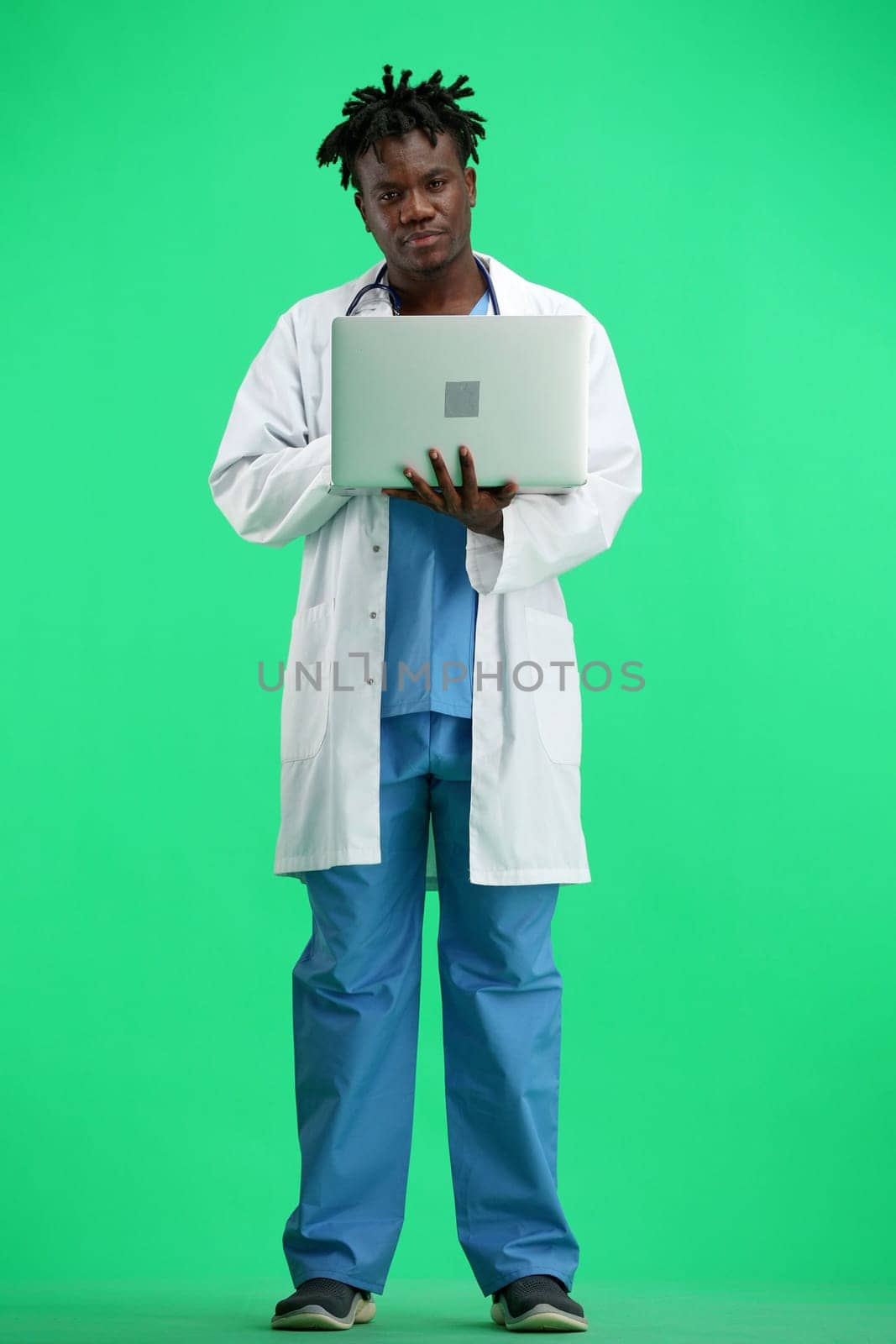The doctor, in full height, on a green background, uses a laptop.