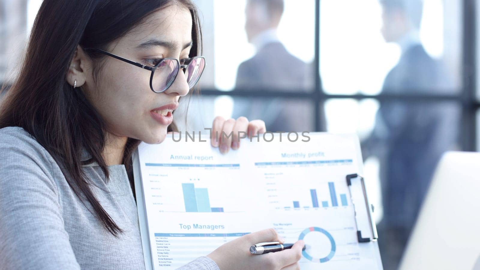 A young woman with glasses in the office is talking on an online connection and showing graphs.