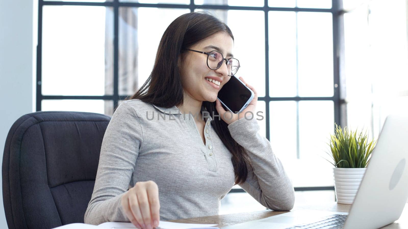 A young woman with glasses in the office is working on a laptop and talking on the phone.