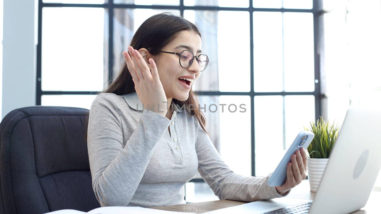 A young woman with glasses in the office is working on a laptop and talking on the phone.