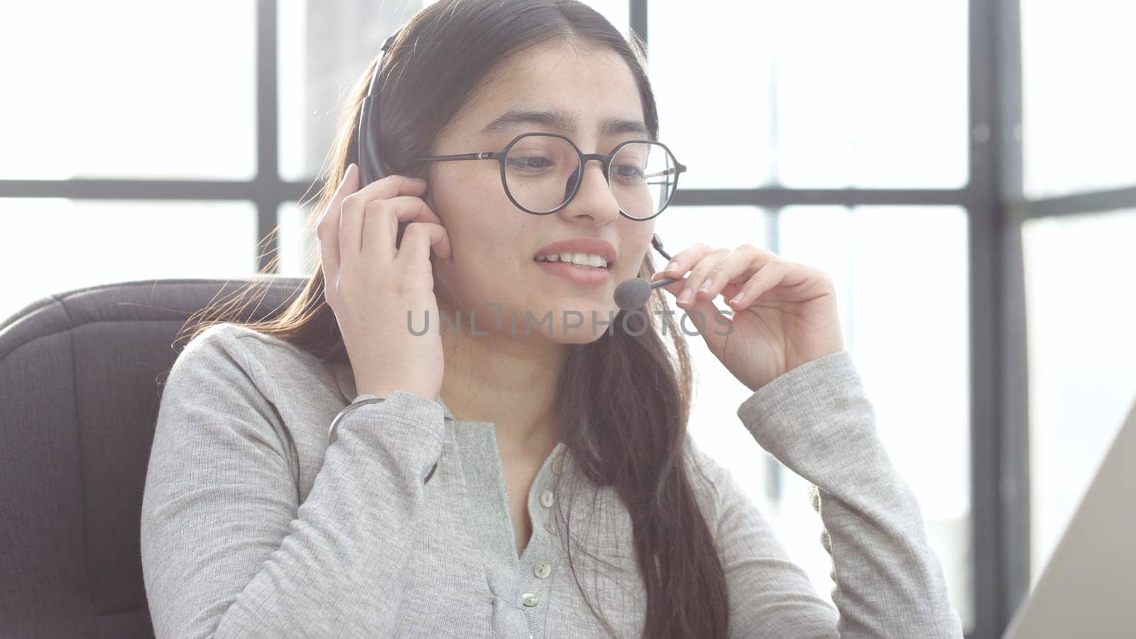 A young woman in close-up with glasses and headphones works in a call center.