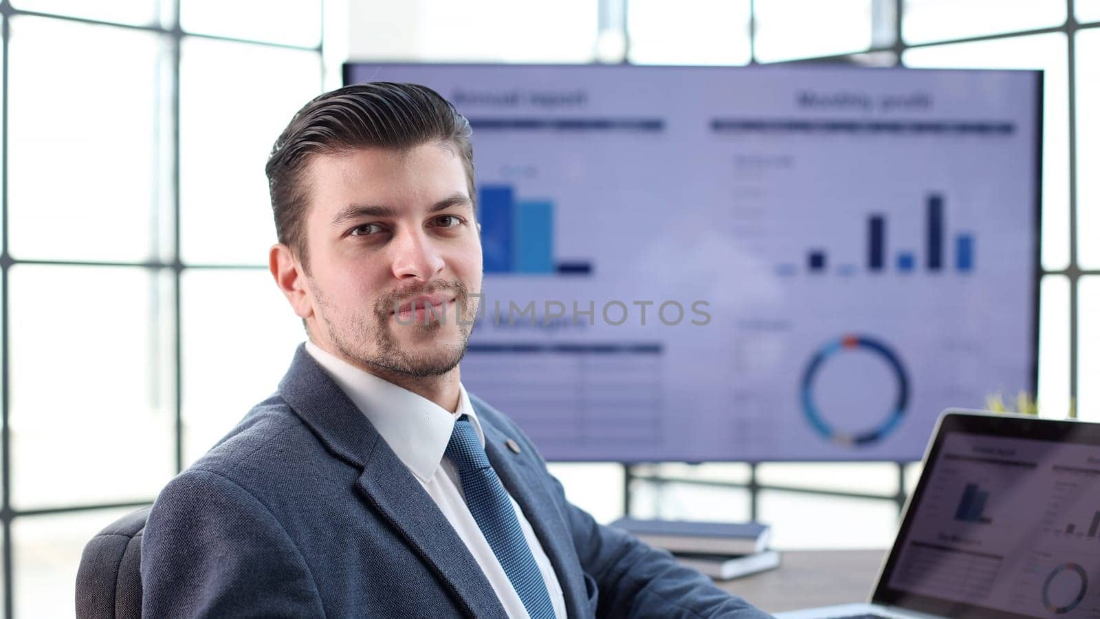A businessman man in a close-up in a jacket with graphs on the background looks at the camera.