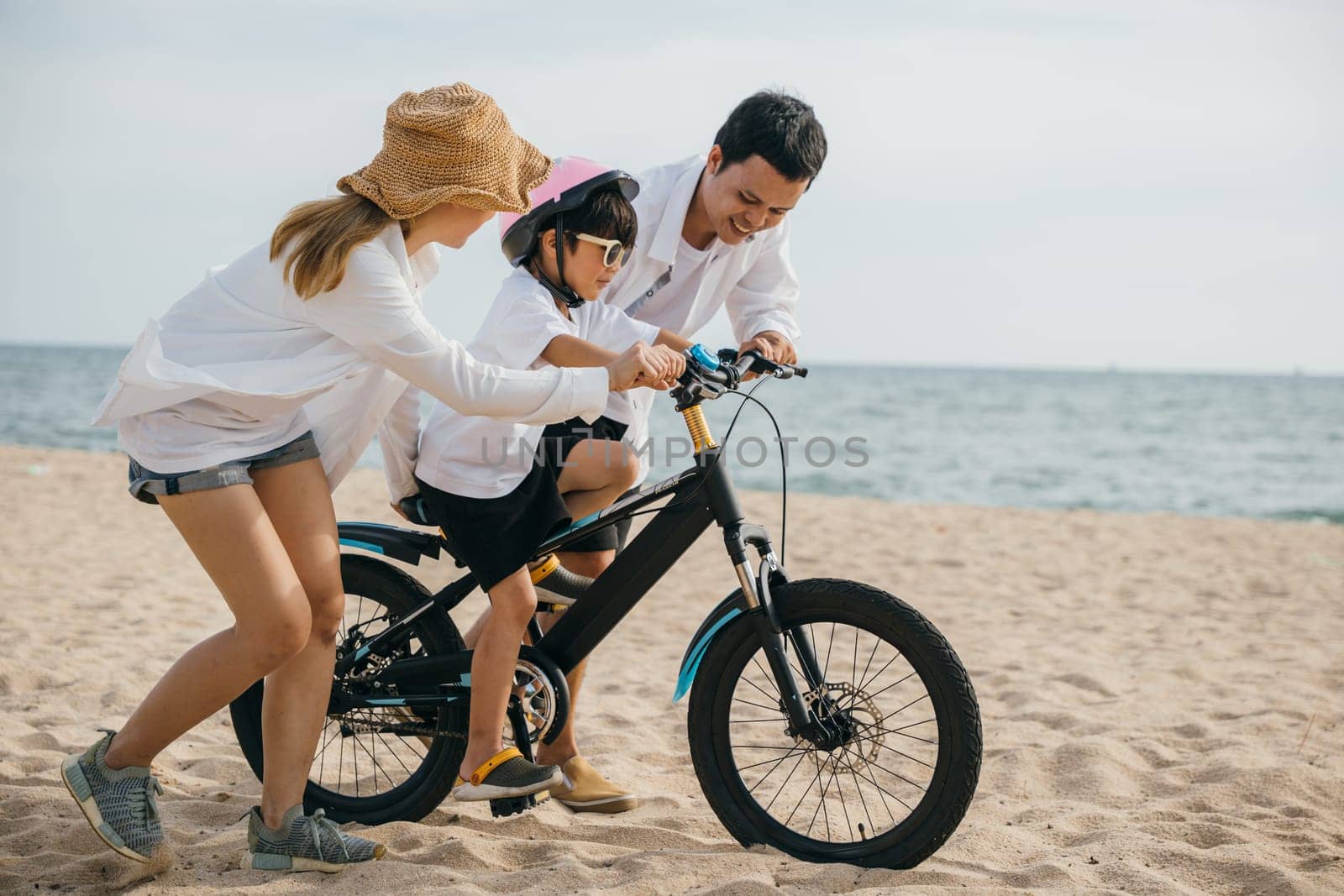 Under warm glow of summer sunset cheerful family enjoys road trip on sandy beach teaching and learning bicycle riding. Safety laughter and family love create perfect vacation concept. by Sorapop