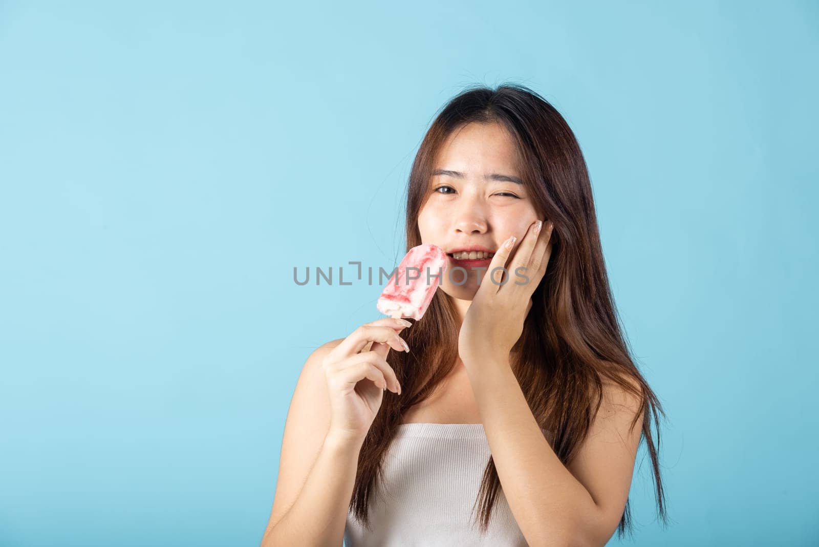 Portrait of Asian young woman with sensitive teeth after eating delicious ice cream wood stick mixed fruit flavor feeling painful uncomfortable, studio shot isolated on blue background, dental problem