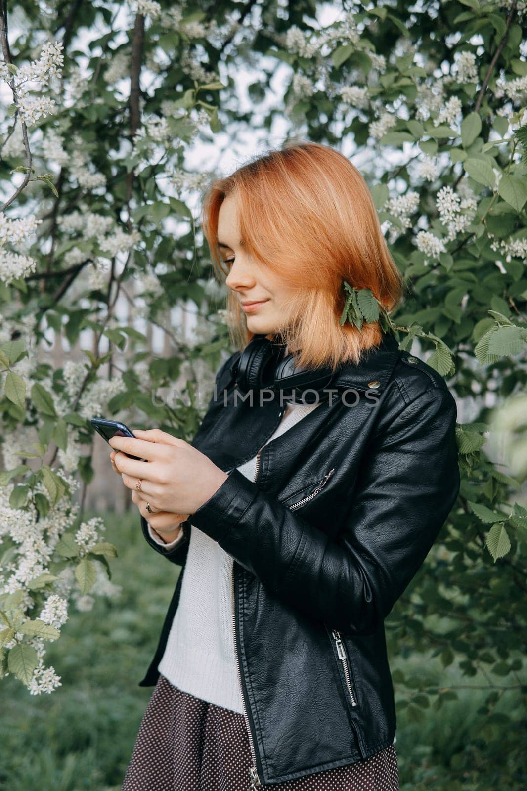 A red-haired teenage girl with headphones around her neck takes a selfie against the background of a flowering cherry tree. The time of spring flowering of cherry orchards