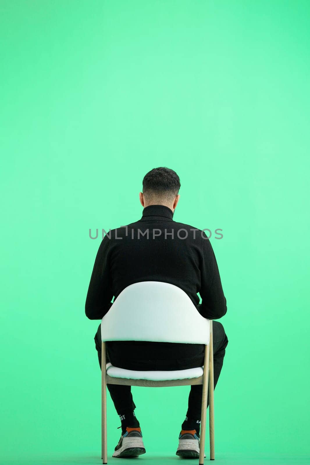 A man, full-length, on a green background, sitting on a chair.