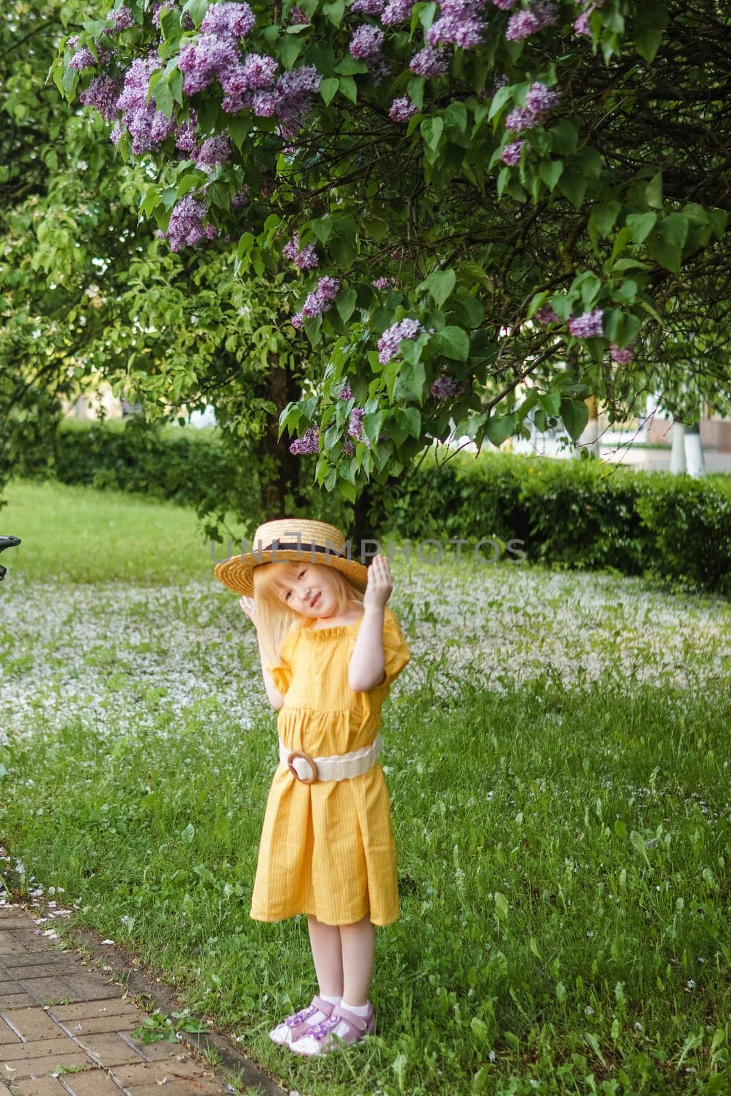 A little girl in a yellow dress and straw hat wearing a bouquet of lilacs. A walk in a spring park, blossoming lilacs