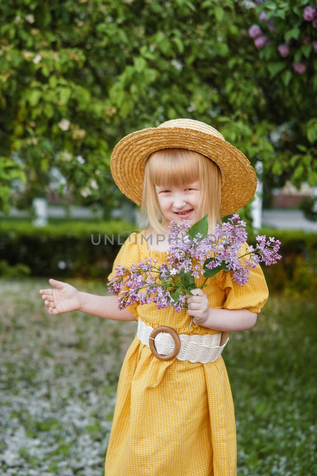 A little girl in a yellow dress and straw hat wearing a bouquet of lilacs. A walk in a spring park, blossoming lilacs