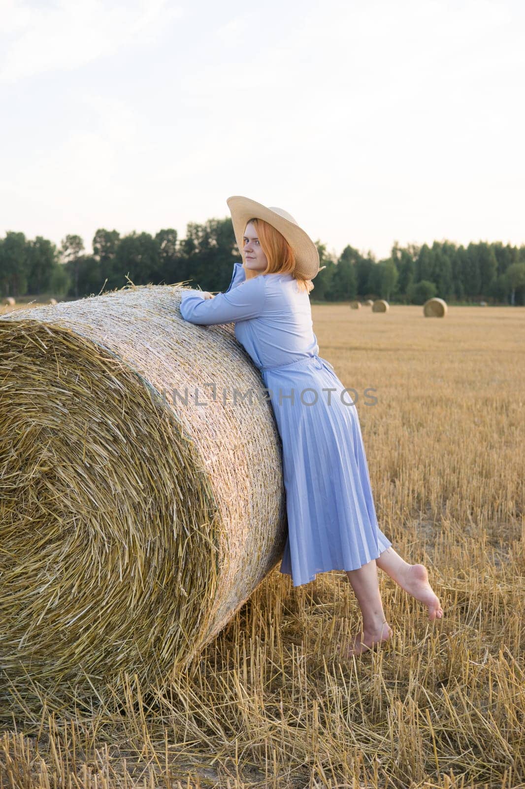 A red-haired woman in a hat and a blue dress walks in a field with haystacks