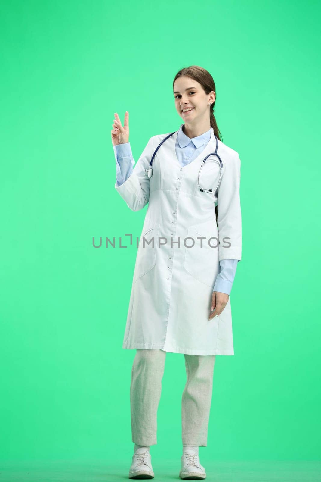 A female doctor, full-length, on a green background, shows a victory sign.