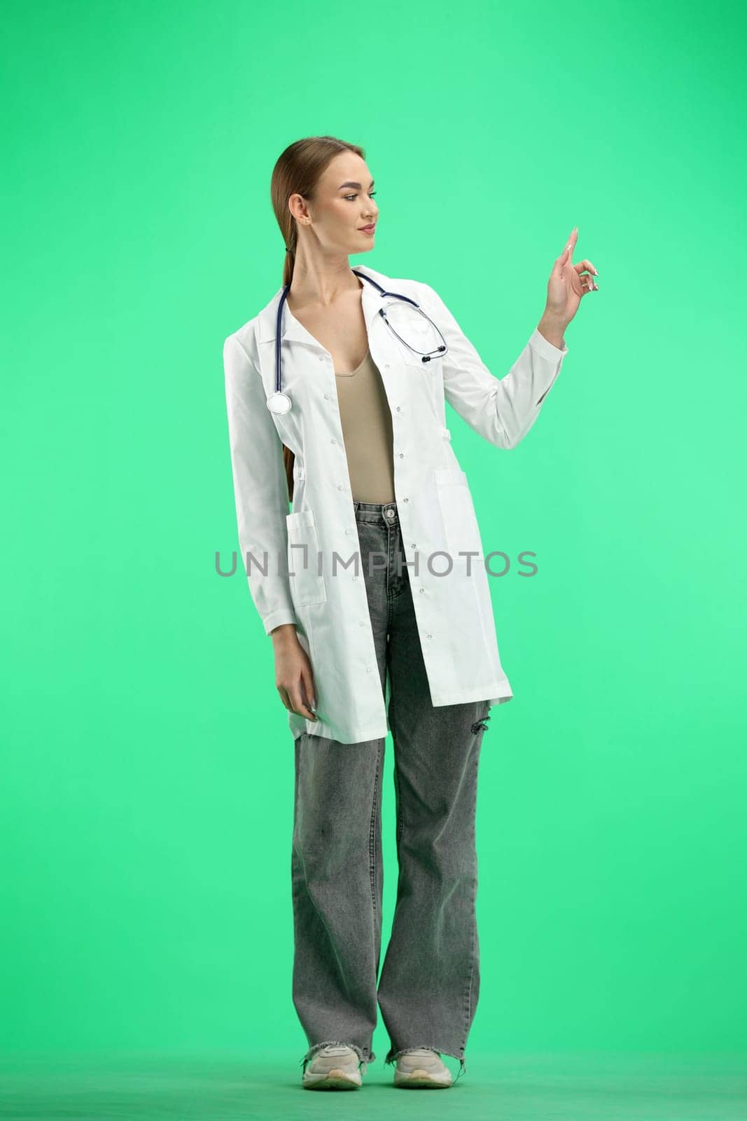 A female doctor, full-length, on a green background, points up.