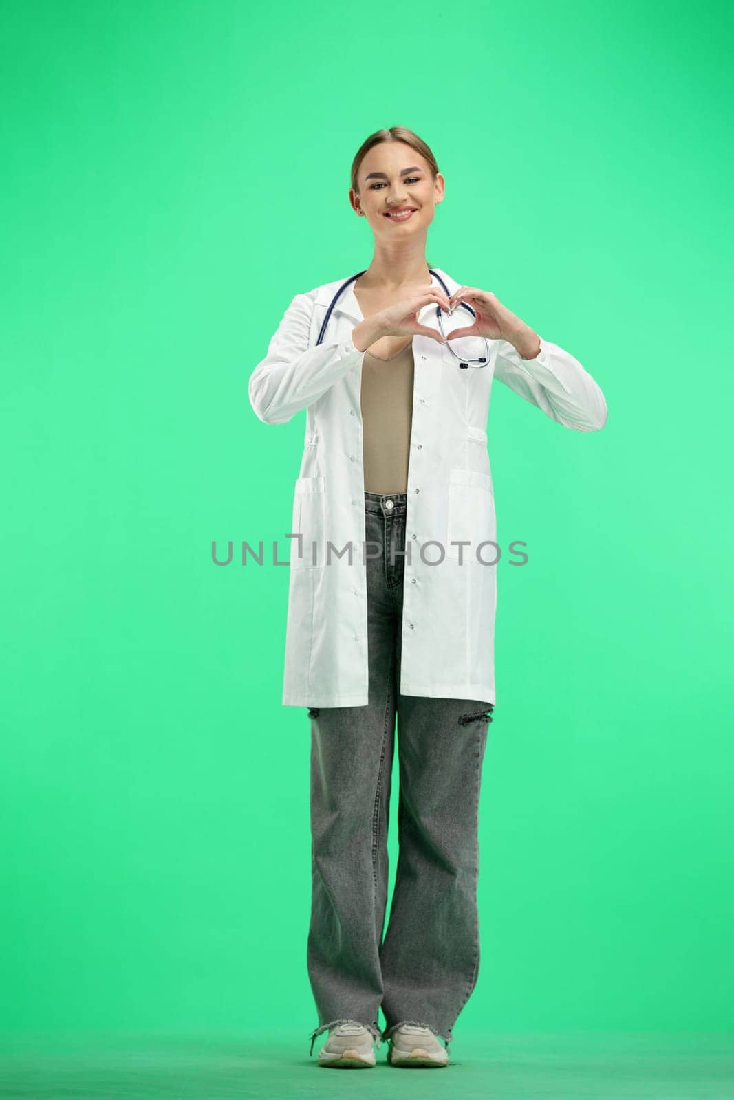 A female doctor, full-length, on a green background, shows a heart.