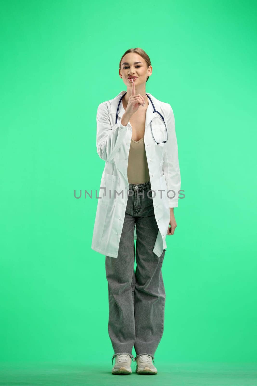 A full-length female doctor, on a green background, shows a sign of silence.