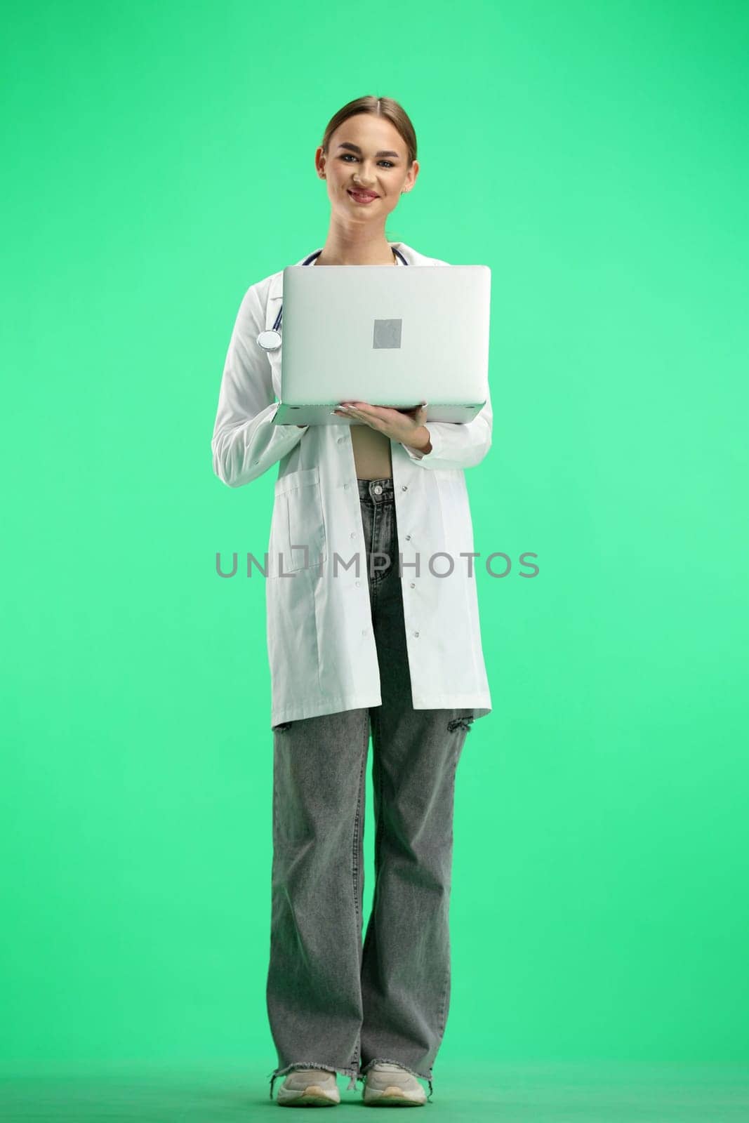 A female doctor, full-length, on a green background, uses a laptop.