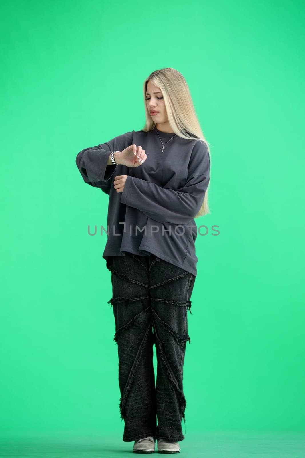 A woman, full-length, on a green background, looks at her watch.
