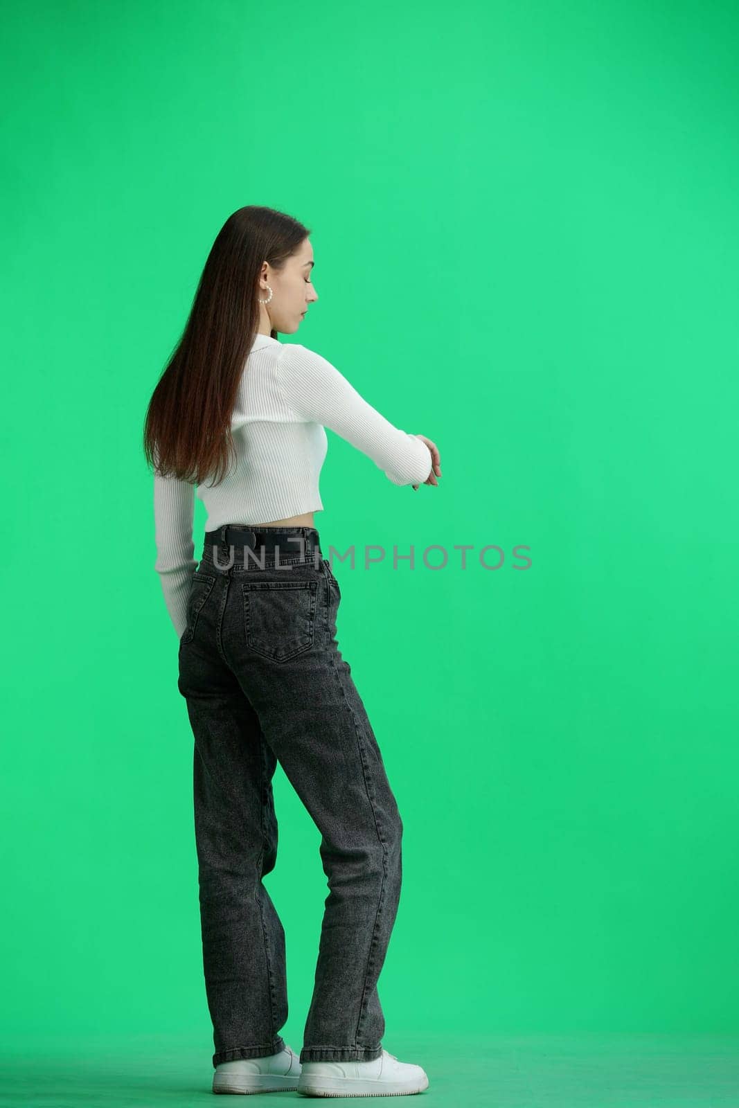 A woman, full-length, on a green background, looks at her watch.