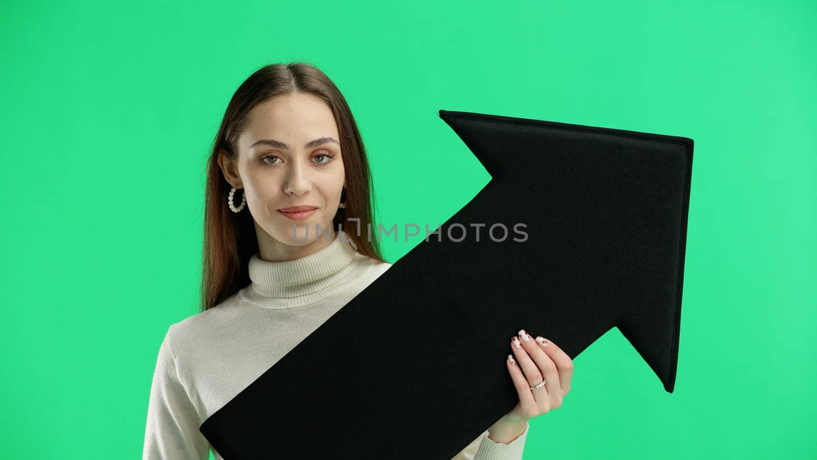 A woman, in close-up, on a green background, points an arrow to the side.