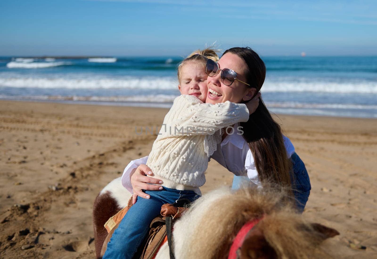 Adorable Caucasian little child girl hugging her mother while riding a horse on the beach by artgf