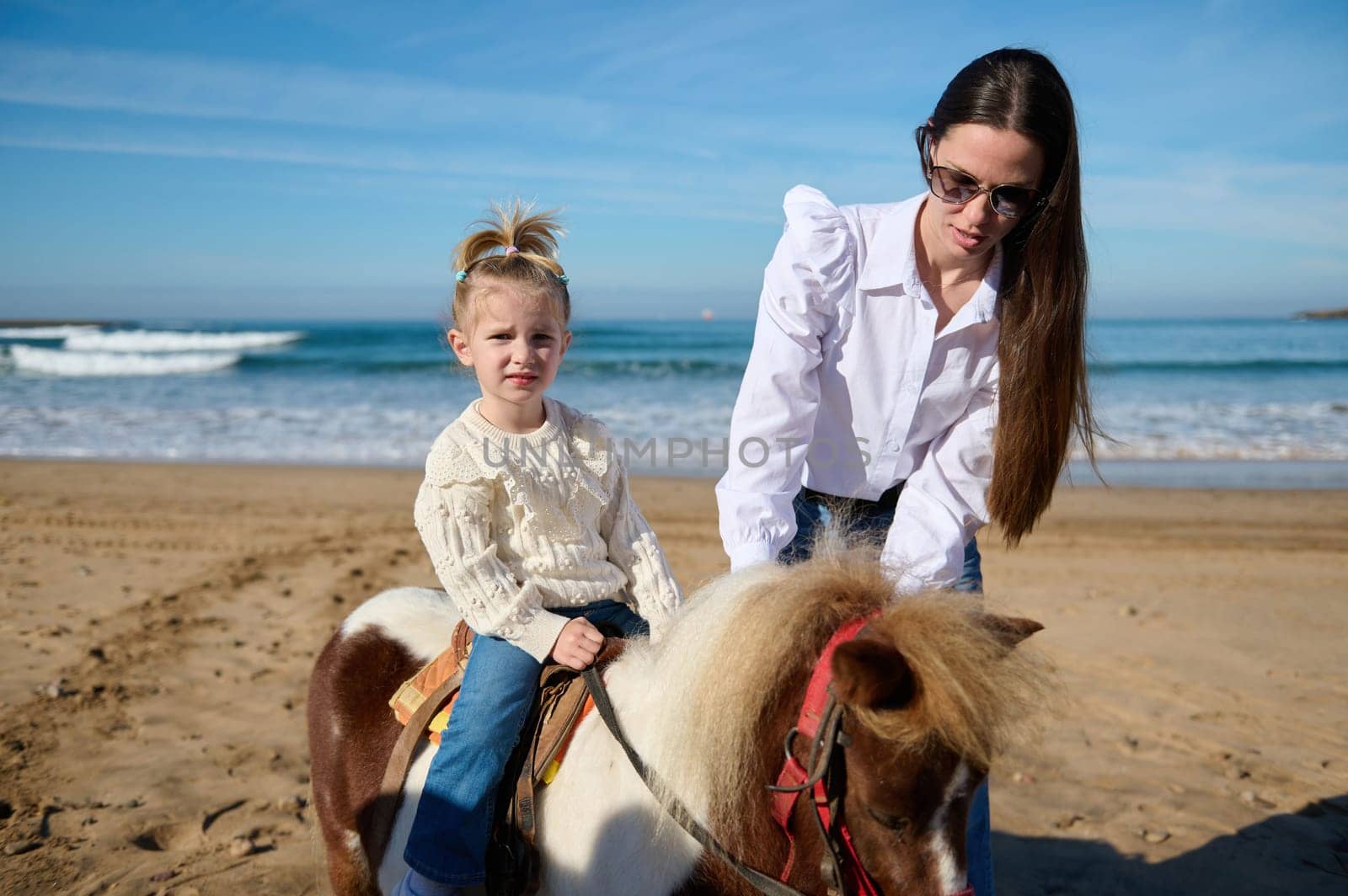 Authentic family enjoying happy time on the sandy beach. Loving caring mother teaching her lovely child to rise a horse pony. People. Nature. Animals. Happy carefree childhood. Leisure and hobbies.
