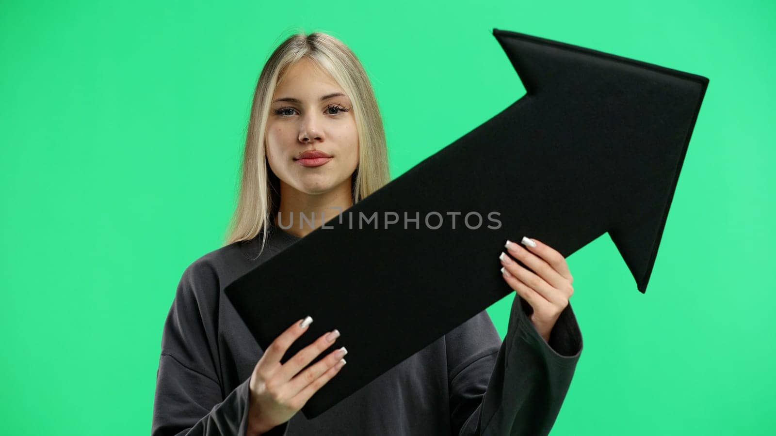 A woman, in close-up, on a green background, points an arrow to the side.