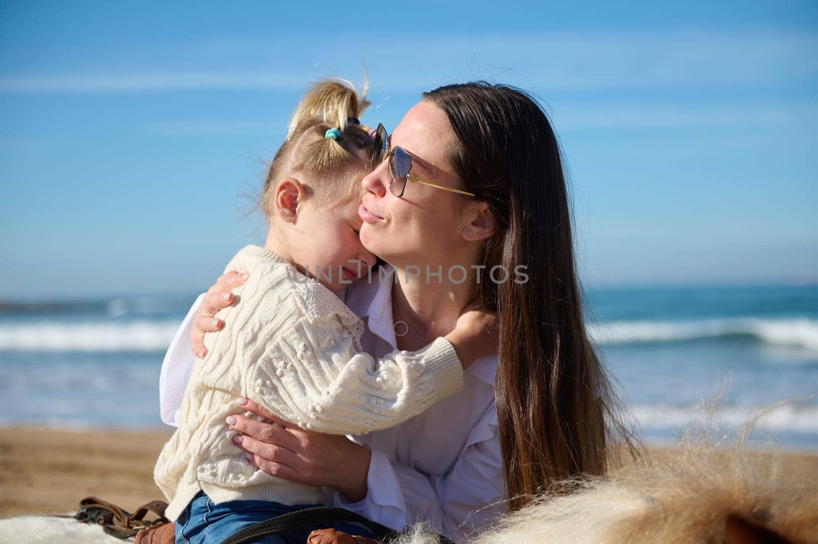 Happy affectionate mother cuddling her little daughter who learns riding a pony outdoor. Happy family relationships. Carefree childhood and maternity leave. People and kindness for animals