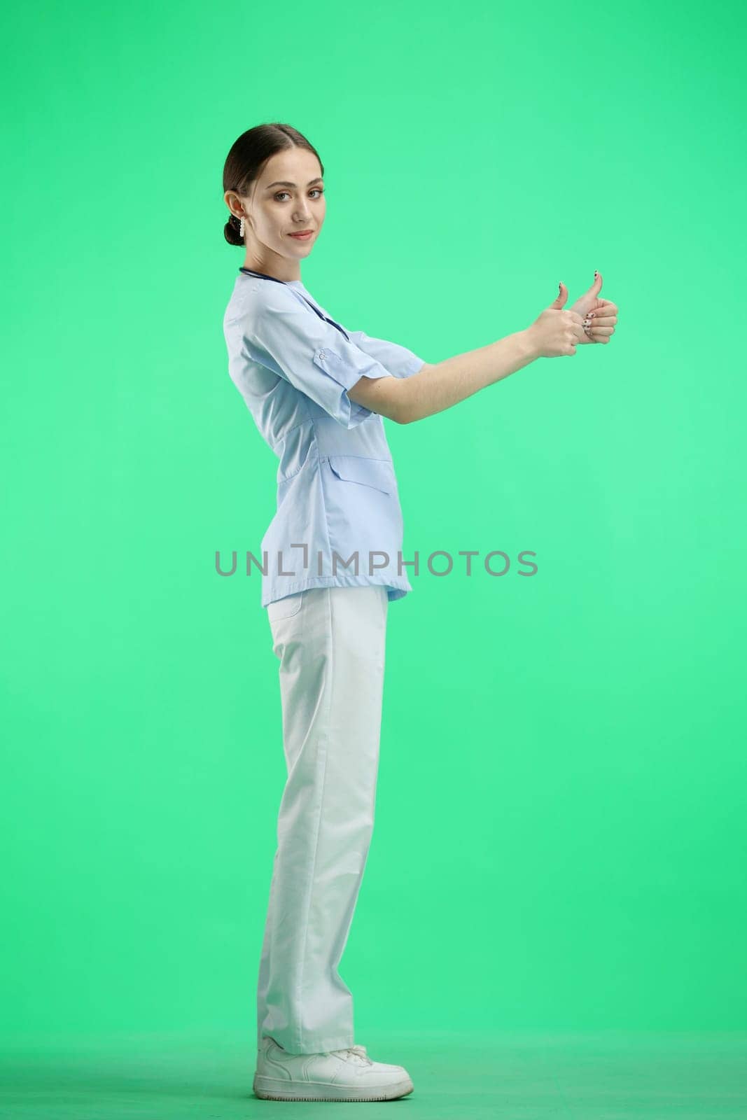 A female doctor, full-length, on a green background, shows a thumbs up.