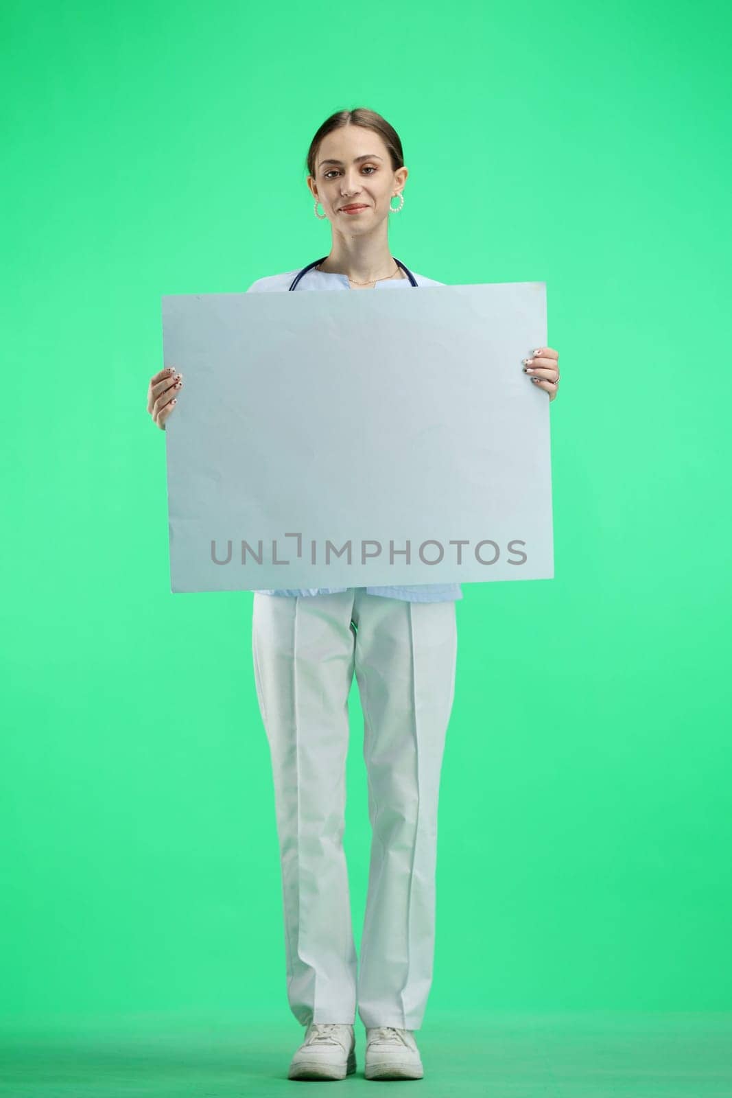 A female doctor, full-length, on a green background, shows a white sheet.