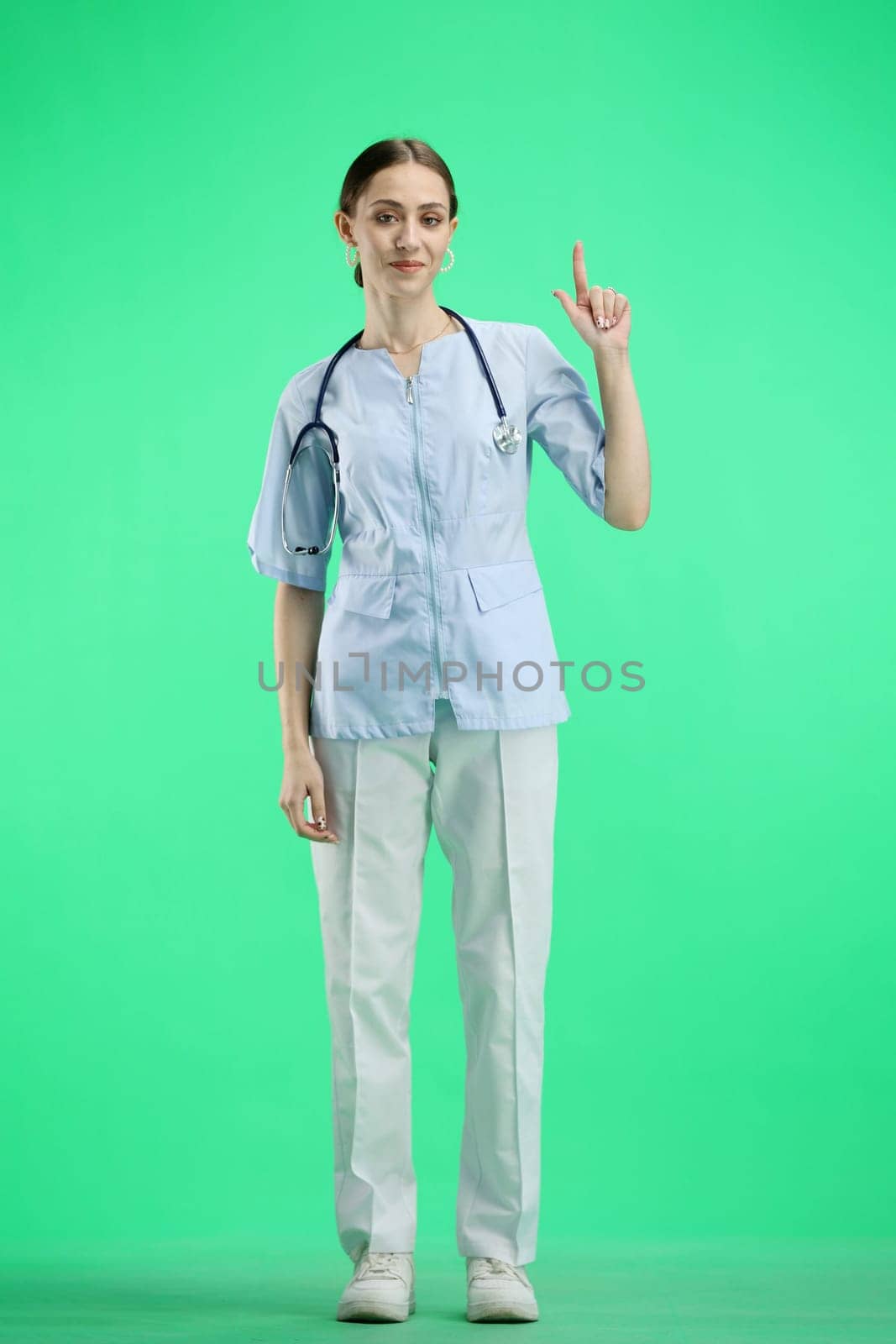 A female doctor, full-length, on a green background, points up.