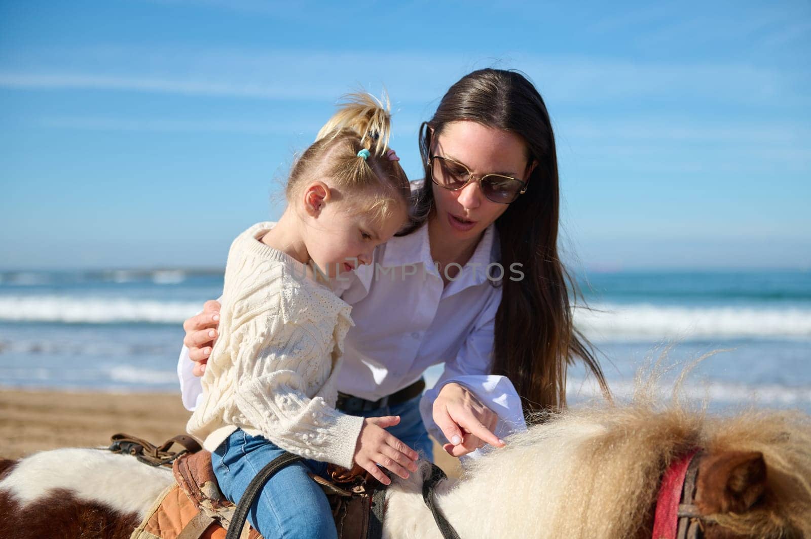 Loving caring mother teaching her daughter to ride a horse pony on the beach by artgf