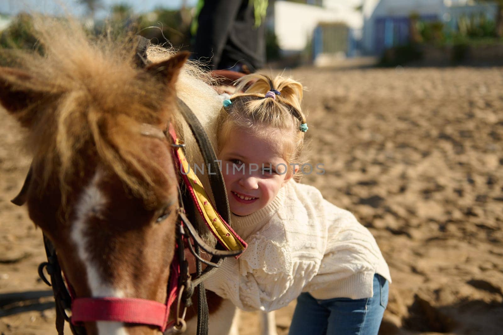 Adorable child girl gently hugging her pony horse, standing on the sandy beach by artgf