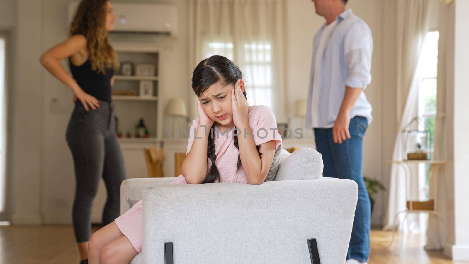 Annoyed and unhappy young girl sitting on sofa trapped in middle of tension by her parent argument in living room. Unhealthy domestic lifestyle and traumatic childhood develop to depression Synchronos