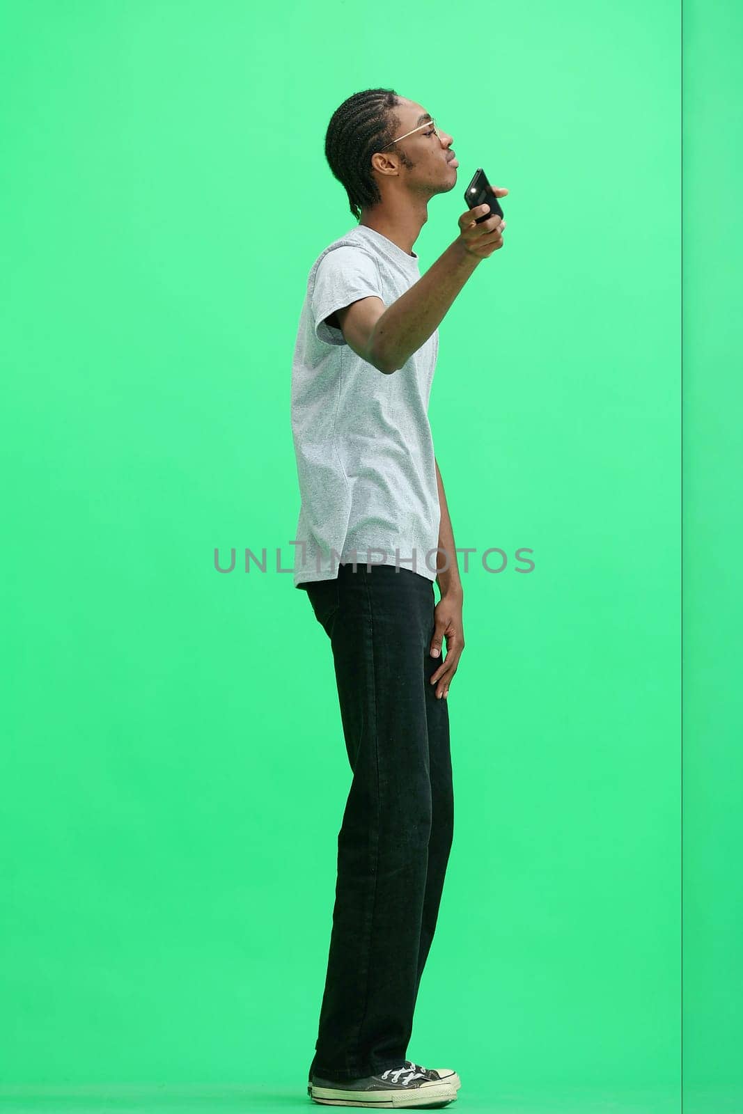 A man in a gray T-shirt, on a green background, in full height, waving his phone.