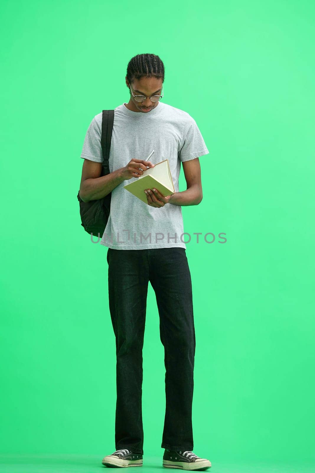 A man in a gray T-shirt, on a green background, full-length, with a backpack, writes.