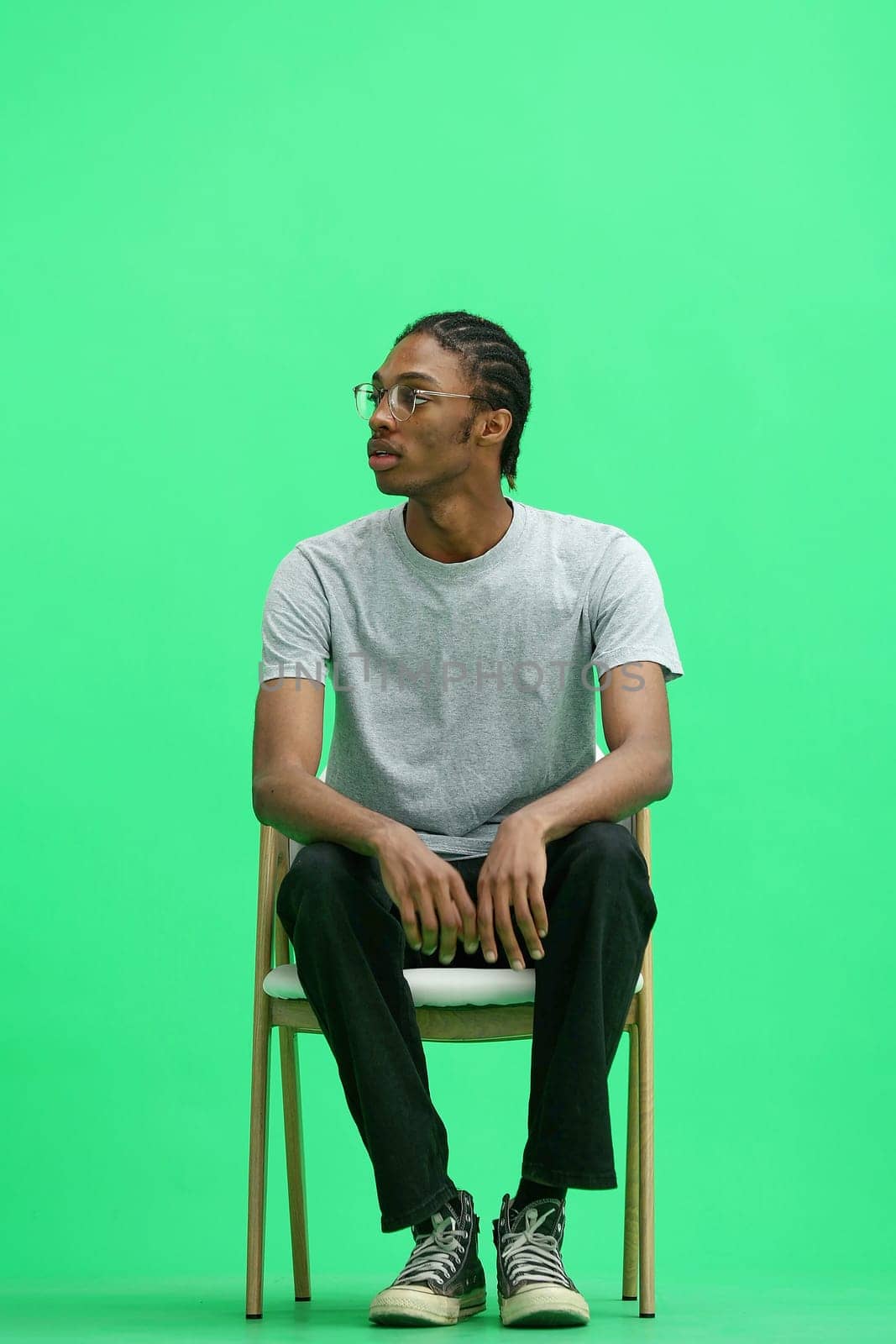 A man in a gray T-shirt, on a green background, full-length, sitting on a chair.