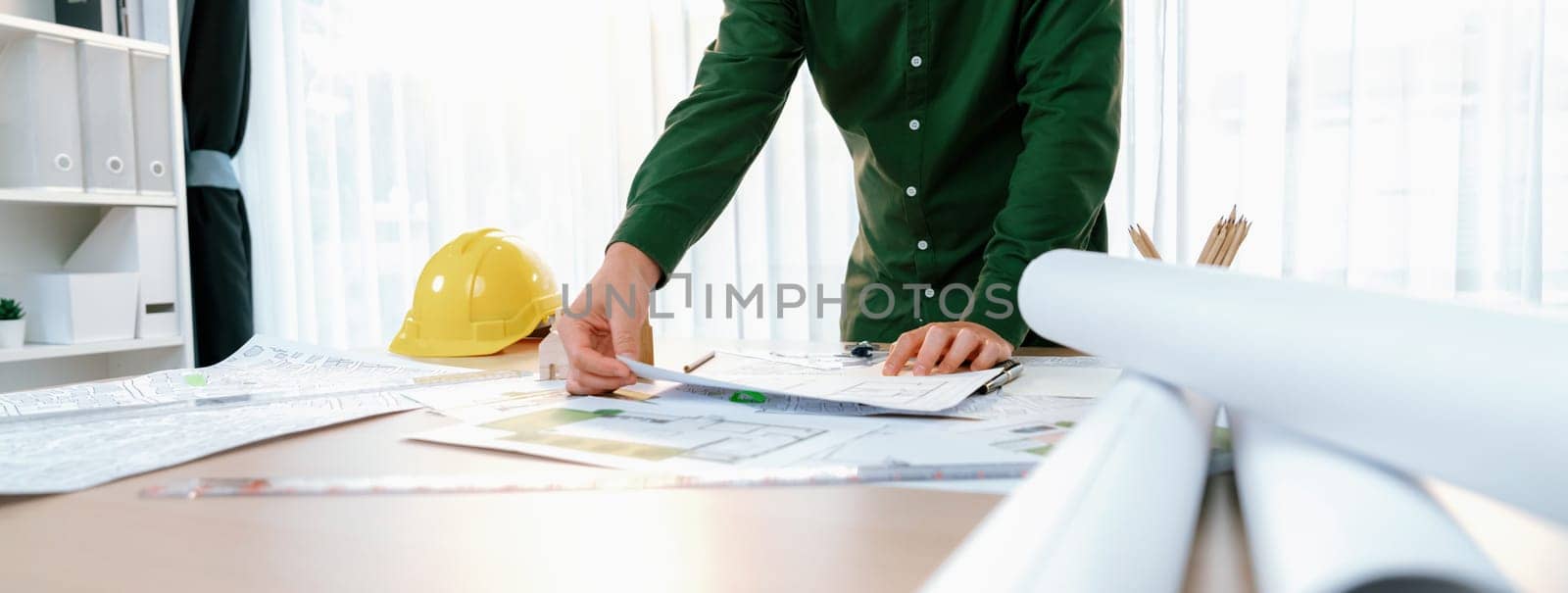 A portrait of professional male engineer shows mistake point in blueprint on messy table with architectural plan and yellow helmet placed at modern office. Closeup. Focus on hand. Delineation.