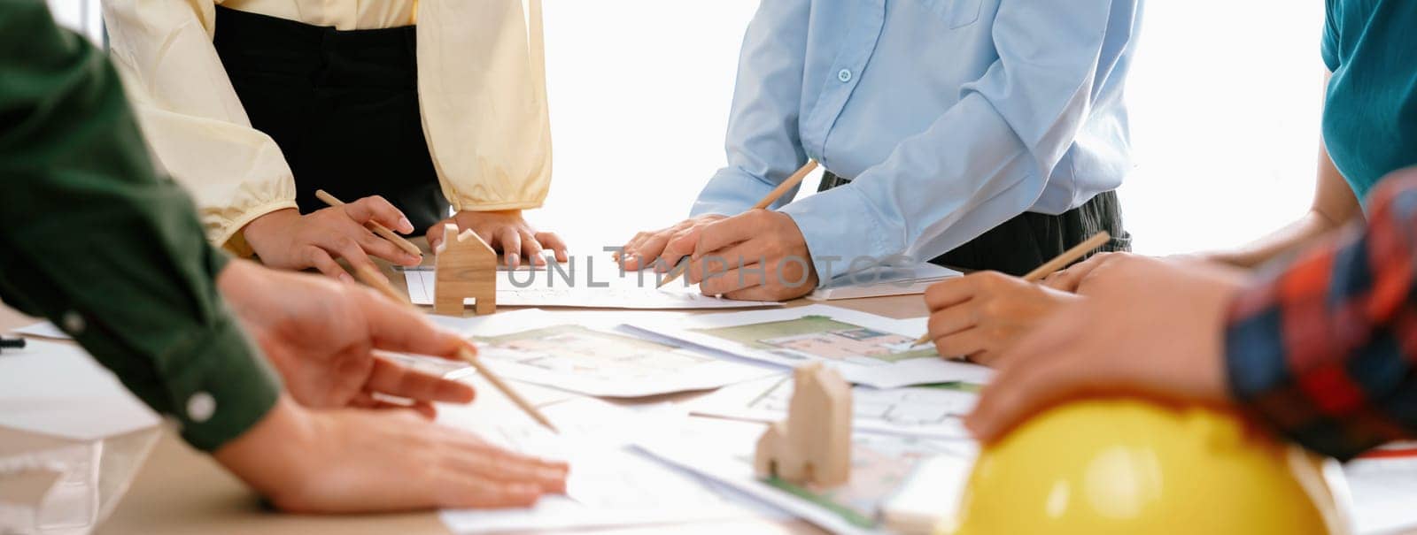 Professional architect team and engineer discuss about architectural project on meeting table with safety helmet, wooden block and architectural document scatter around. Closeup. Delineation.