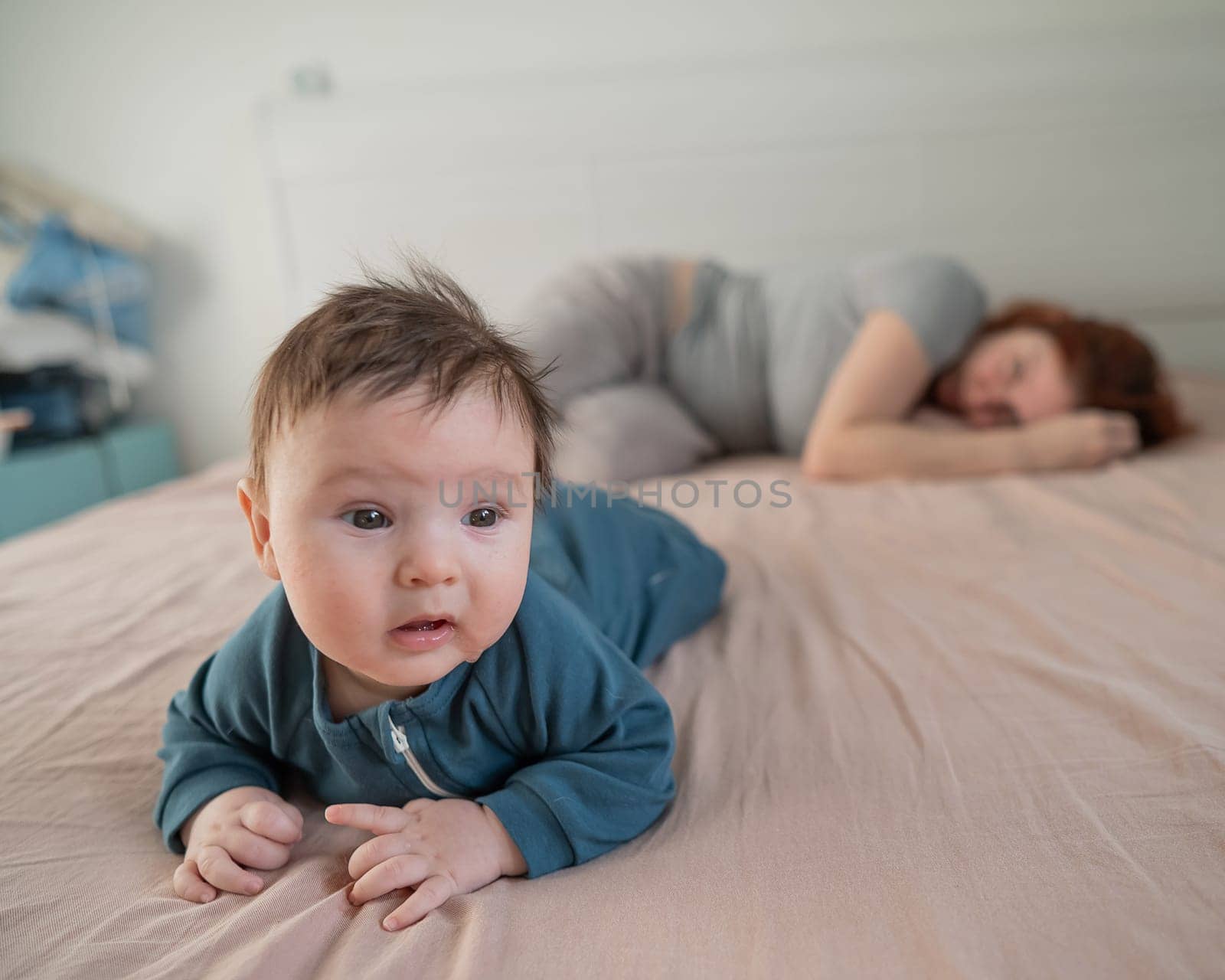 A three-month-old boy lies on his stomach on the bed and his mother sleeps behind him. Postpartum depression