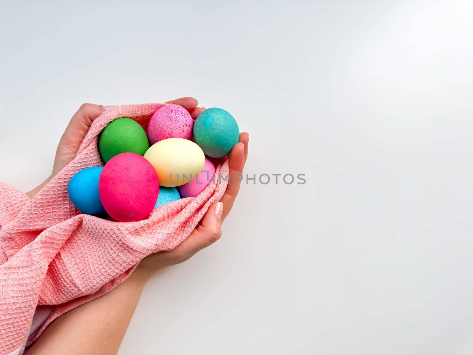 Hands tenderly hold variety of colorful Easter eggs wrapped in soft pink fabric on white background with empty space for text. Suitable for seasonal celebrations and festive spring looks. by Lunnica