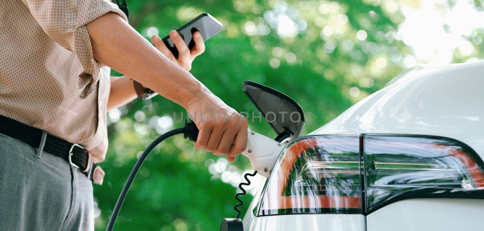 Young man use smartphone to pay for electricity at public EV car charging station green city park. Modern environmental and sustainable urban lifestyle with EV vehicle. Panorama Expedient