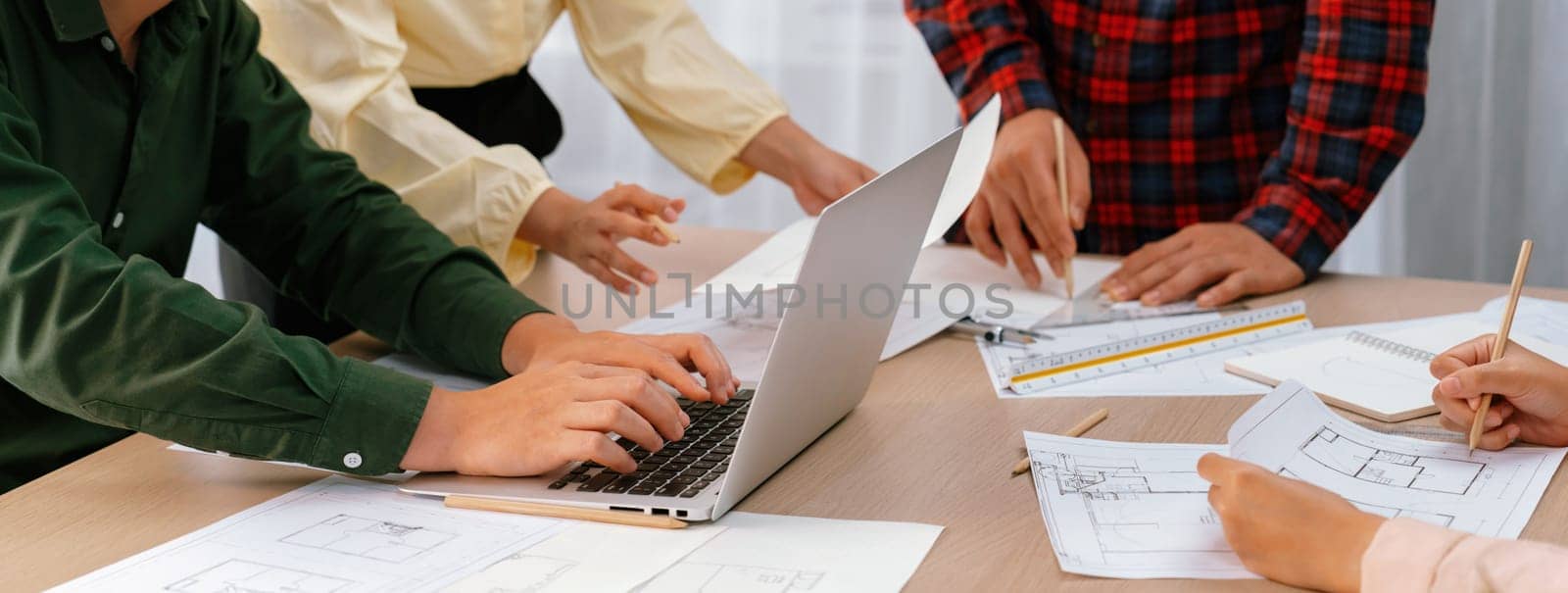 Expert engineer team discussion about construction design while male engineer using laptop analysis data on table with blueprint and architectural equipment scatter around. Closeup. Delineation.