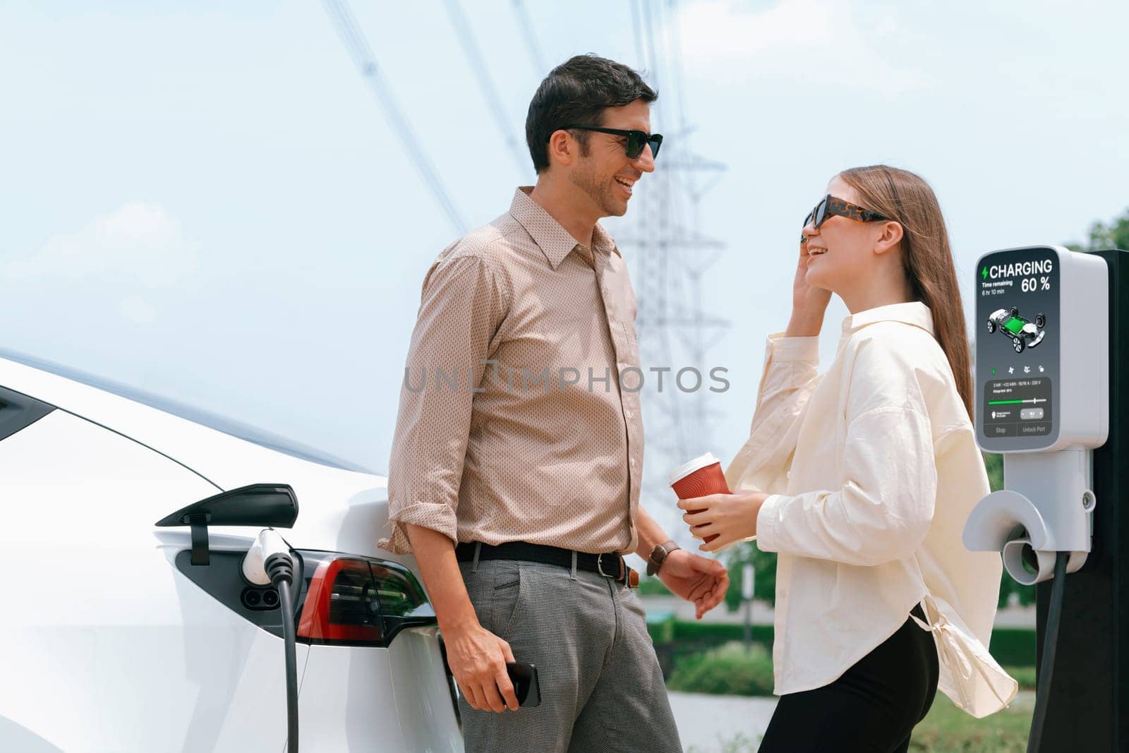 Young couple with coffee recharge EV car battery at charging station connected to electrical industrial power grid tower. Couple with shopping and travel using eco electric car lifestyle. Expedient