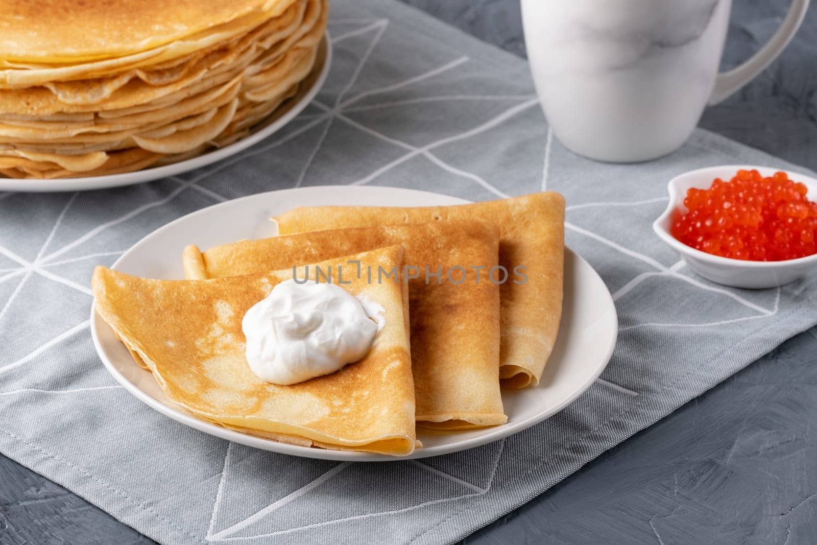 Pancakes with sour cream and red caviar. Close-up of pancakes stacked in a plate on a gray background.