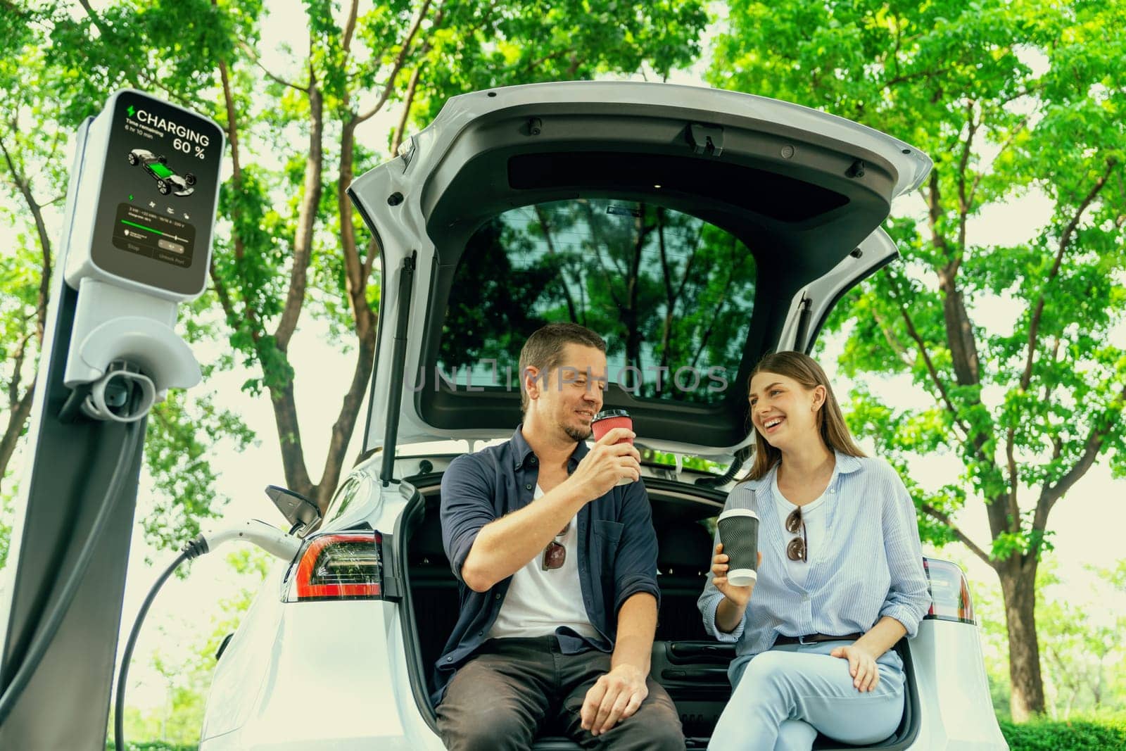 Lovely young couple drinking coffee while recharging battery for electric car during road trip travel EV car in natural forest or national park. Eco friendly travel during vacation and holiday. Exalt