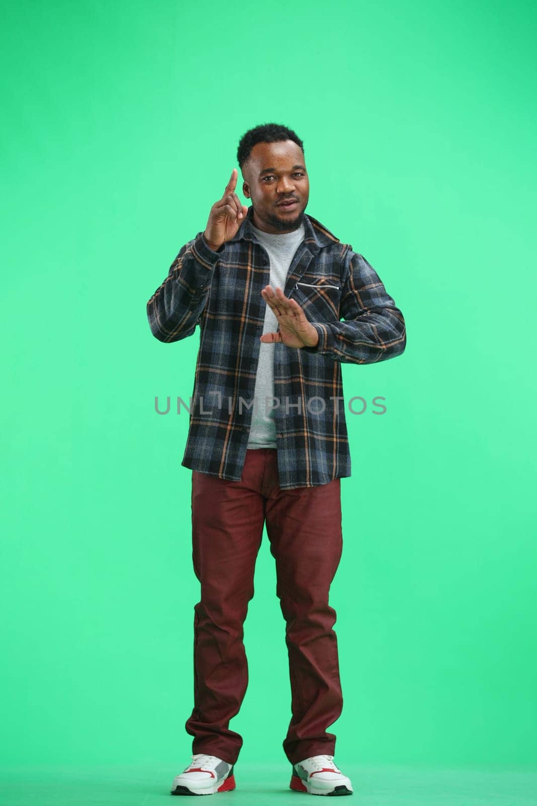 A man, full-length, on a green background, gestures.