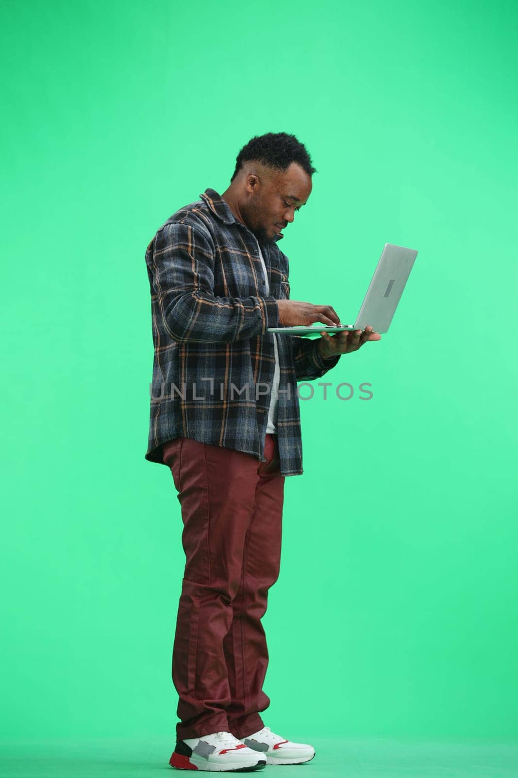 A man, full-length, on a green background, uses a laptop.
