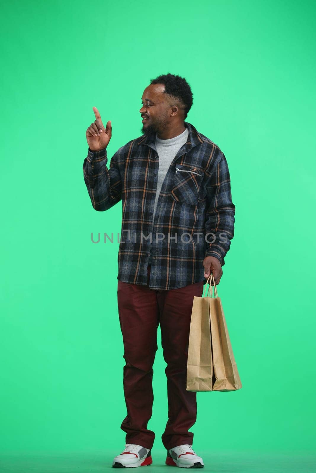 A man, full-length, on a green background, with bags, points up.