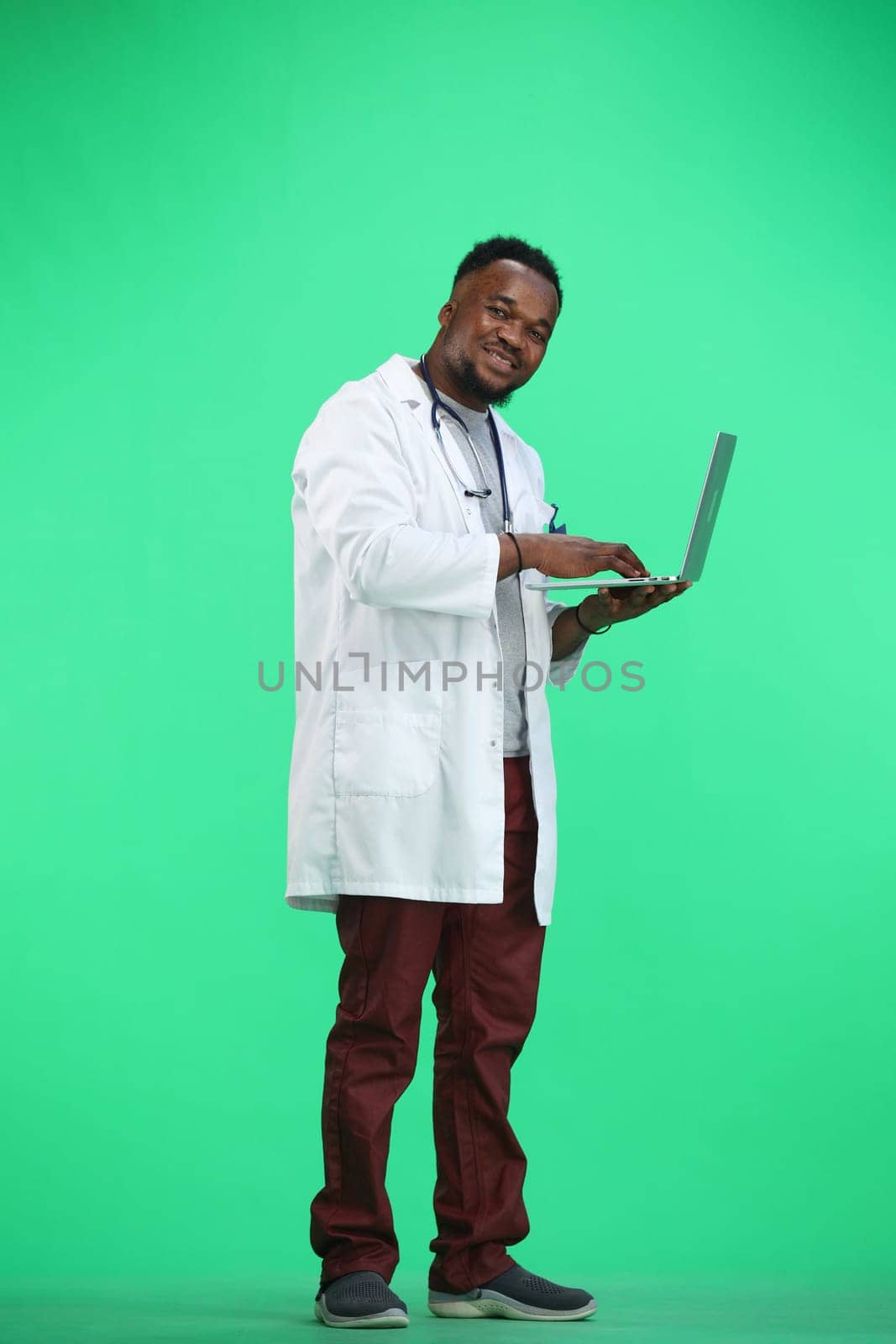 The doctor, in full height, on a green background, uses a laptop.